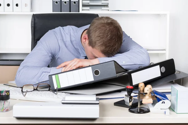 young exhausted businessman lying on the desk in the office in front of a stack of files. in the background there is a shelf. the man looks down.