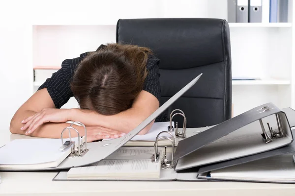 一个年轻的 灰心丧气的女人躺在办公桌前的一堆文件前 后面是一个架子 — 图库照片
