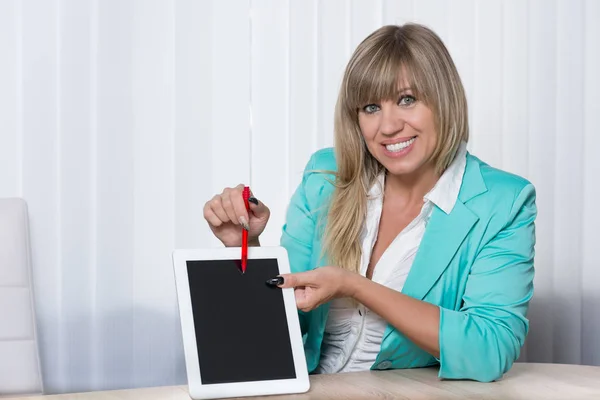 a woman sitting at table in office and pointing with a pen on a tablet. the woman looking to the camera.