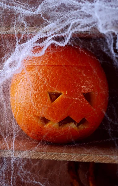 Spooky spider web covered jack-o-lantern formed from a fresh orange on a wooden shelf in a scary Halloween background