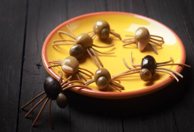 Scary Halloween spider appetizers made with cured black and green olives with spaghetti legs crawling over a yellow plate on a buffet against a dark background clipart