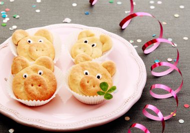 Delicious cookies forming small pigs with small green clover leaves on pink plate. Served on the table with gray cloth cover, colorful sequence and spiral thin wrapping foil. clipart