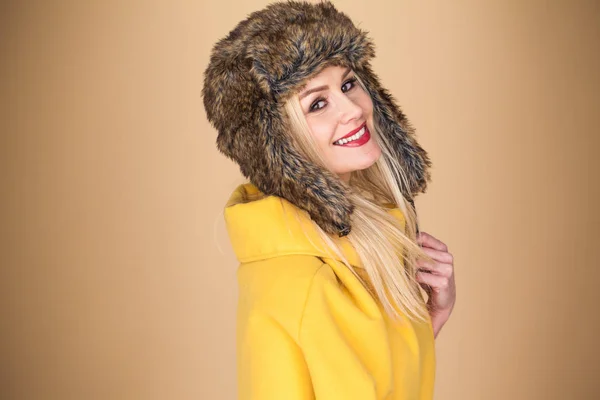 漂亮的金发女人 有着可爱的活泼的笑容 头戴时髦的冬季毛皮帽 背景为棕色 头戴典雅的黄色上衣 — 图库照片