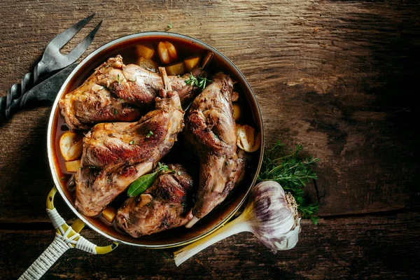High Angle View of Roasted Rabbit Haunches in Pan with Stewed Vegetables on Rustic Wooden Table Surface with Bulb of Garlic and Copy Space