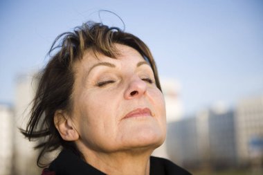 side head portrait of a mature woman in front blurred urban background with closed eyes a deep breath clipart