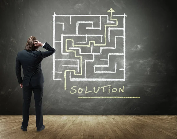 Rear View of Business Person Scratching Head with Hand in Confusion Standing in front of Chalkboard with Drawing of Maze with Solution in Business Concept Image