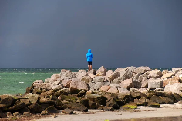 man stands alone on a stone beach by the sea while a storm front approaches.