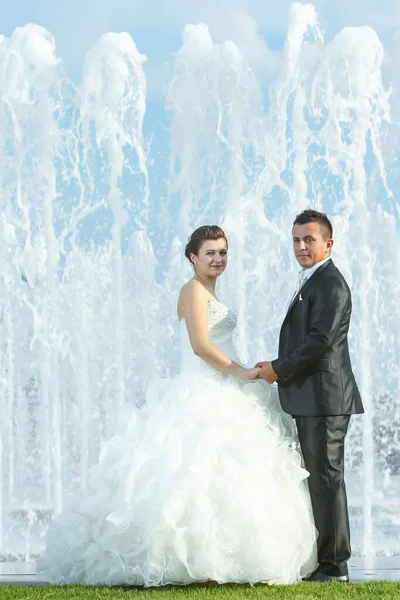 The bride and groom holding hands and looking at camera while standing in front of a water spray fountain in Zagreb, Croatia.