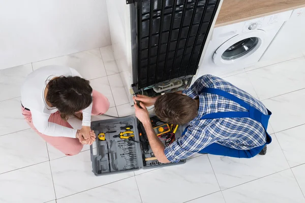 Young Repairman Repairing Refrigerator In Front Of Beautiful Woman