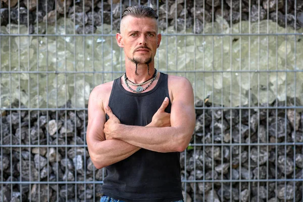 Tough guy with sparrow beard, undercut and black rip shirt standing thoughtful in front of a wall of stones