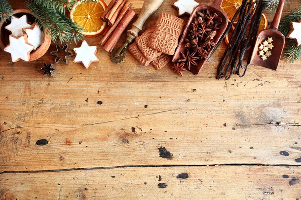 Traditional Christmas spices and cookies arranged as a top border over rustic wood background with cinnamon, star anise, speculoos and star biscuits and dried orange slices, large copyspace
