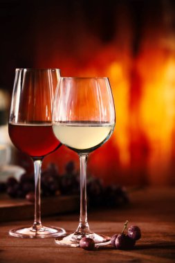 Two glasses of red and white wine on a wooden table in front of a blazing fire with selective focus to the glasses of beverage clipart