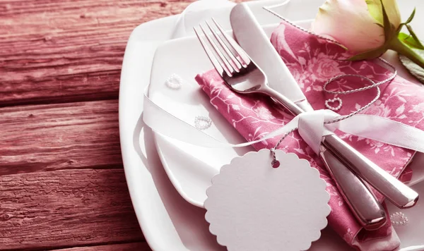 Blank Tag Tied Around Silver Knife and Fork Resting on Pink Napkin on top of White Dishes and Decorated with White Rose and Small Pearl Hearts - Romantic Wedding Place Setting with Copy Space