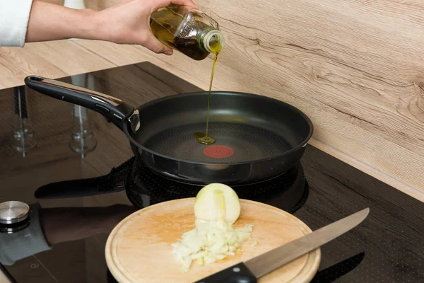 Pouring oil in the pan for cooking, onion with knife on cutting plate in the foreground