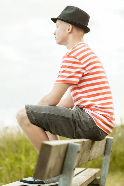 Side view of calm male teenager in hat, striped shirt and shorts sitting on top of wooden bench near wild grass