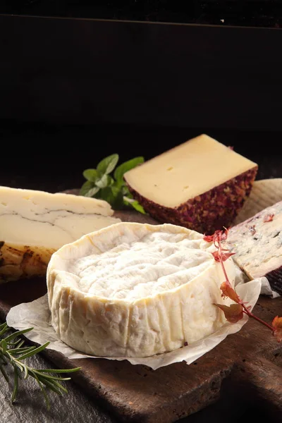 木製ボード上で提供される柔らかい高齢者のチーズのラウンド他の様々なグルメチーズとコピースペースと黒の背景の前に新鮮なハーブで飾られた — ストック写真