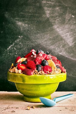 Scrumptious acai berry bowl of cereal overflowing with blueberries, slices of bananas, strawberry and coconut shreds next to blue spoon on old wooden table clipart