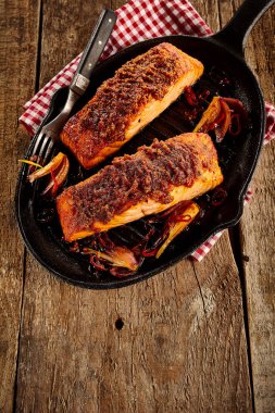 Spicy baked salmon with seasoning and fork in black cast iron griddle over napkin and rustic wooden table surface clipart