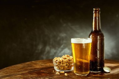 Beer served from a brown bottle into a long glass and a bowl of roasted salted peanuts on an old wooden bar table with copy space on slate behind clipart
