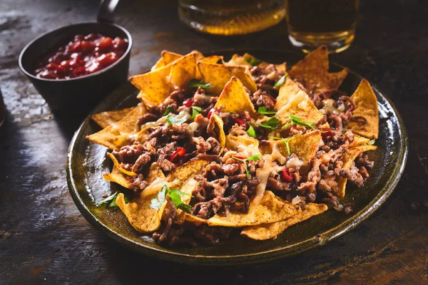 Tortilla chips garnished with ground beef, melted cheese, peppers and cilantro leaves in plate on wooden table