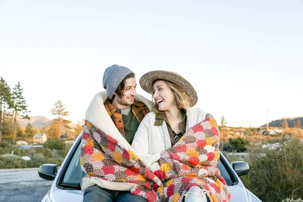 Young couple on car bonnet wrapped in patchwork blanket face to face smiling, Chilao Campgrounds, Los Angeles, California, USA
