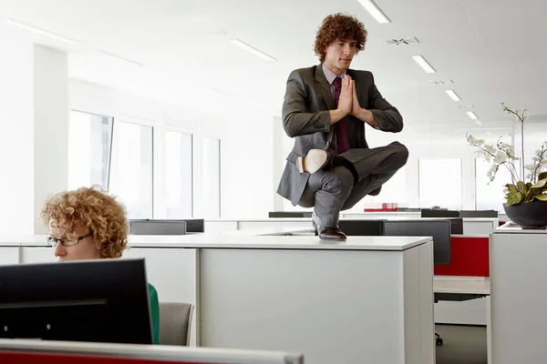 Businessman doing yoga pose on top of filing cabinet, behind woman working at desk