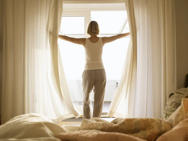 パジャマを着た熟女が寝室のカーテンを開ける — ストック写真