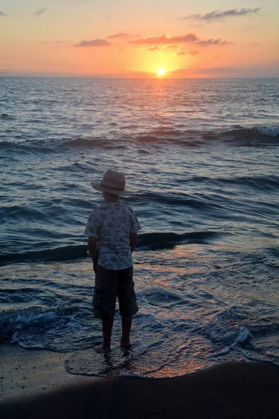 小さな男の子が夕日のビーチに立っていてズボンのポケットに手を入れて地平線を見ています — ストック写真