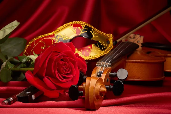 Violin (fiddle), theater mask and red rose lying on the perfect red satin fabric. String instrument.