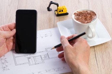 Close-up of architect hands working on a blueprint and holding a smartphone. Cup of coffee and miniature extractor in the background. Blueprint was created by photographer. clipart
