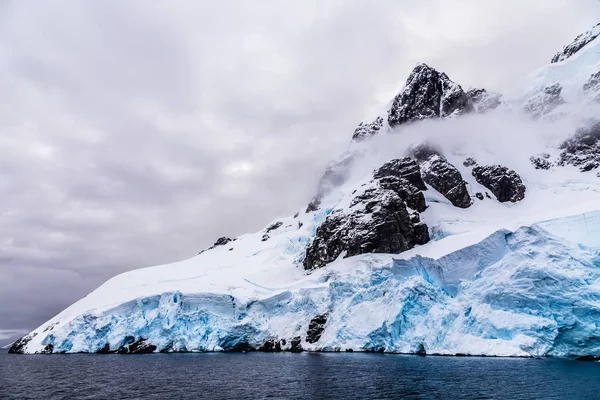 アルゼンチンの島々 南極クルーズの近くで 前景に水で覆われた青い氷河と雲で覆われた巨大な急な石の岩 — ストック写真