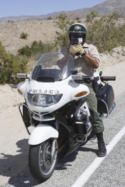 Full length of a mature traffic cop on motorbike monitoring speed though radar gun clipart