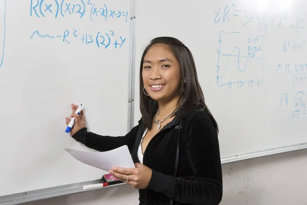 Portrait of happy young female student holding marker and paper in front of whiteboard