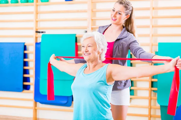 Senior woman with stretch band in fitness gym being coached by personal trainer