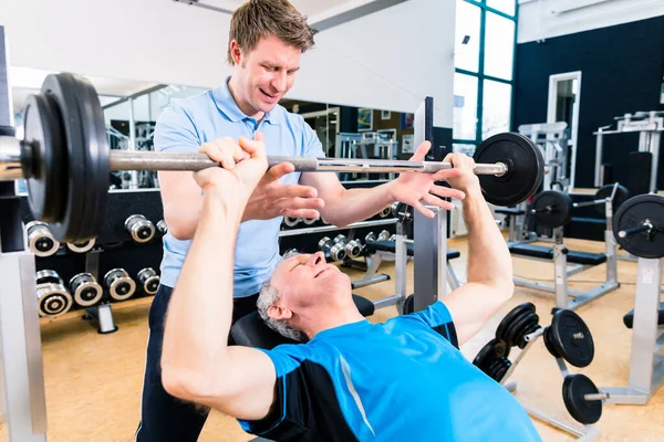 Trainer assisting senior man lifting barbell in gym to gain strength and fitness