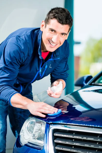 Happy young man looking at camera while waxing a blue car outdoors at car wash