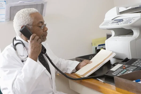 An African American senior male doctor holding document while using landline phone