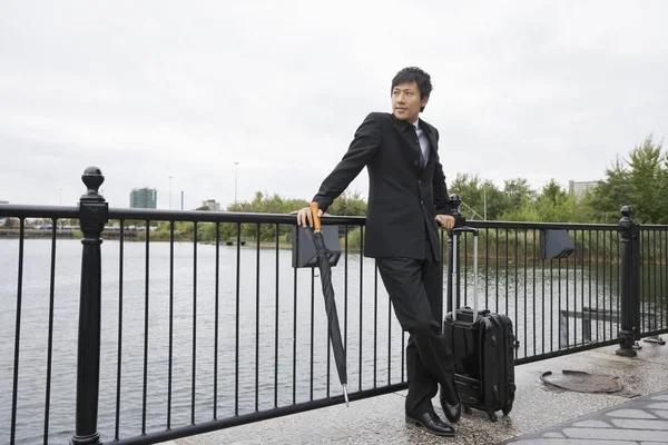 Full length of businessman with luggage leaning on railing along river