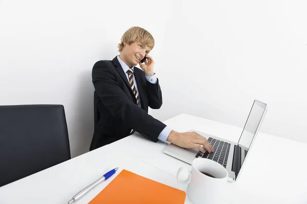 オフィスのデスクでノートパソコンを使用している間に電話でビジネスマンを自信を持って — ストック写真