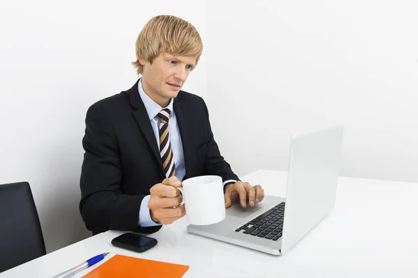 オフィスのデスクでノートパソコンを使いながらコーヒーカップを持っているビジネスマン — ストック写真