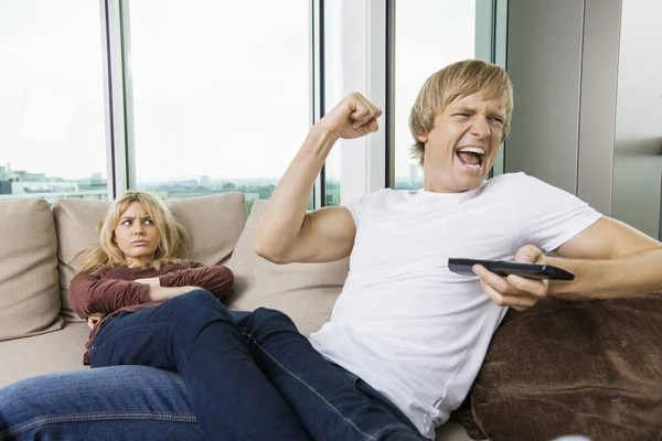 Angry woman staring at cheerful man as he watches TV in living room at home
