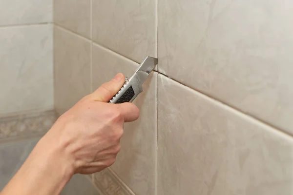 Male hands with knife removing the old grout. Replacing old grout between tiles. Raking out tiles for regrouting.