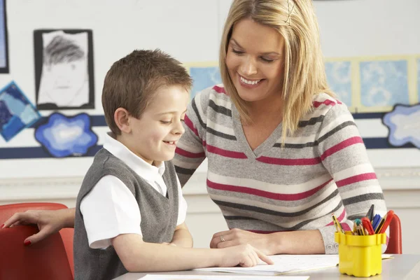 Male Primary School Pupil And Teacher Working At Desk In Classroom