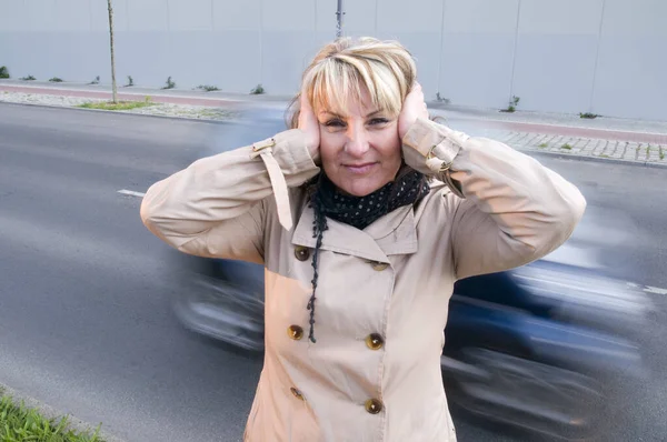 head-and-shoulders shot of a mature woman in a trench coat on the roadside with blurred passing cars in the background,talking to the palms ears.