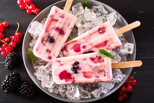 Three healthy frozen yogurt and berry iced lollies or popsicles in a bowl of ice chilling on a party buffet for a tasty summer dessert over black with fresh blackberries and red currants