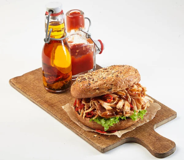 Delicious spicy pulled chicken sandwich or burger on a wholegrain bun served on a wooden board with bottles of ketchup and olive oil dressings isolated on white