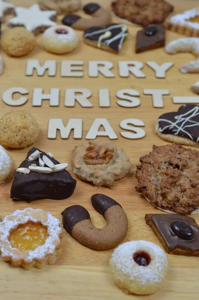 バニラクレセントやマカロンやマジパンのハートのようなクリスマスクッキーは木の文字から出てくる言葉の周りのブレッドボード上の別の種類 メリークリスマス — ストック写真