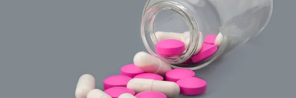 白い錠剤とピンクの錠剤は 灰色の背景にガラス瓶から散乱されます — ストック写真