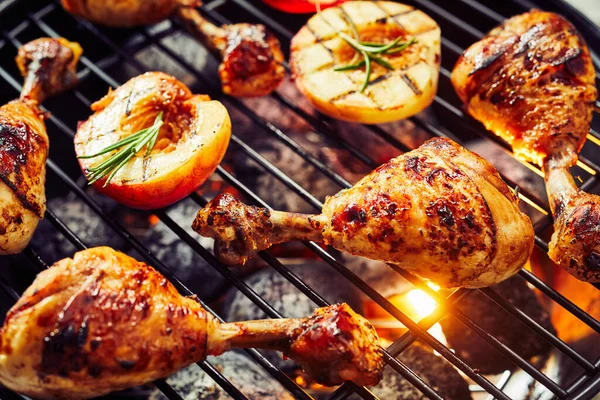 Spicy chicken legs sizzling over a hot barbecue with halved fresh apricots or nectarines seasoned with rosemary in a close up view