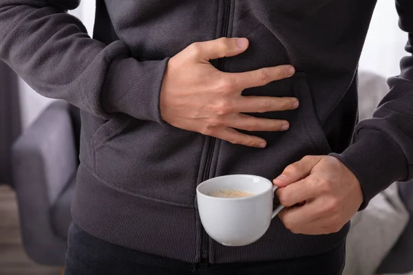 Man Suffering From Stomach Pain With Cup Of Coffee On Wooden Desk